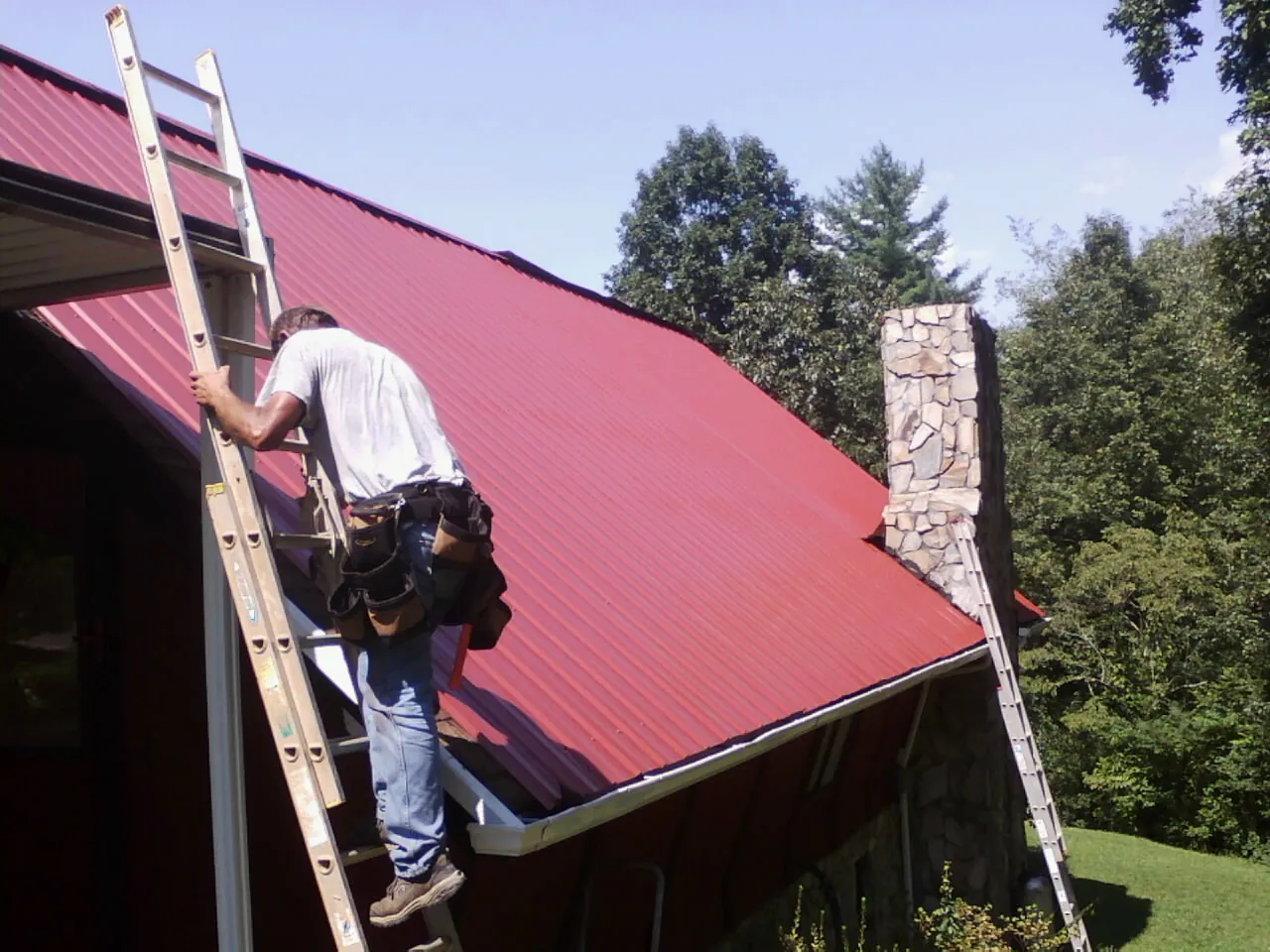 Roofer on a ladder beside a red metal roof and stone chimney — Soffit & Fascia Repair by Roofing Brothers in Rising Sun